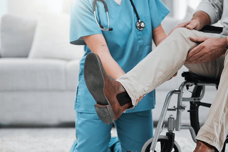 at-home nurse helping an older man in a wheelchair move his leg