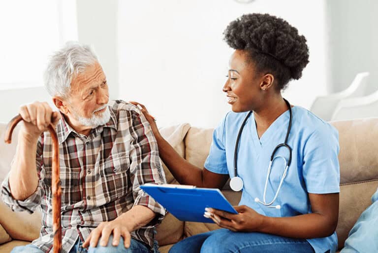 female nurse with a clipboard sitting on a couch talking with a senior man