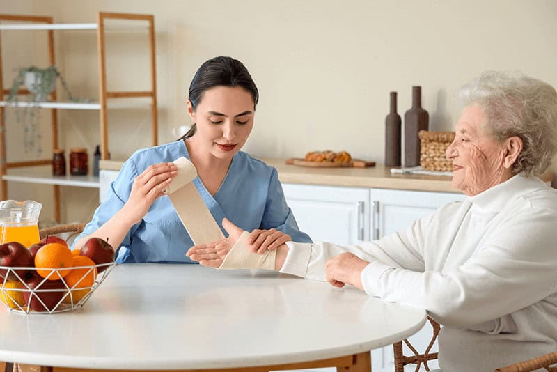 in-home nurse bandaging a senior woman’s wrist at a dining room table